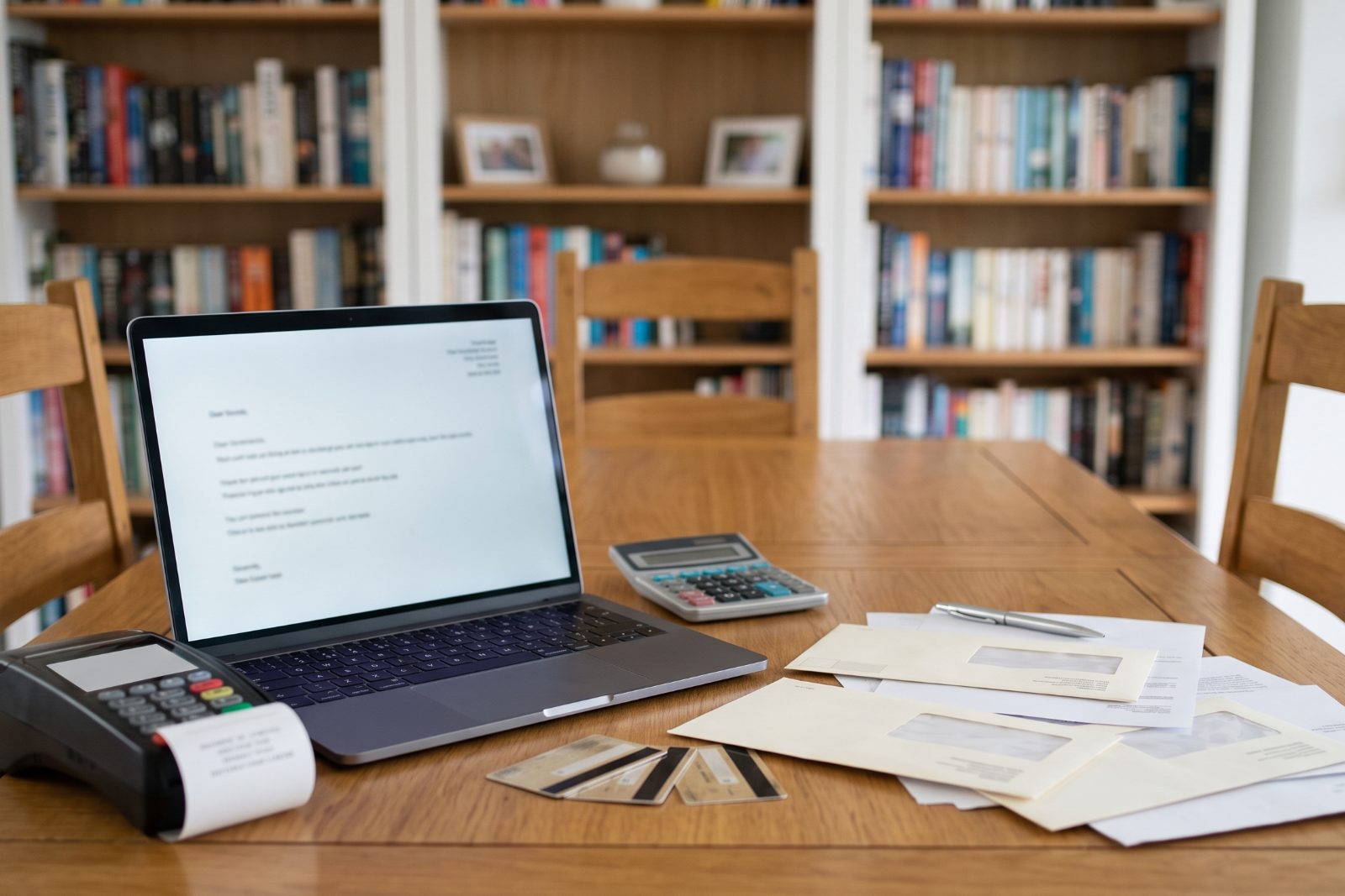 A dining table at home with a laptop, a calculator, a card terminal, bank cards and opened letters, in front of full bookshelves